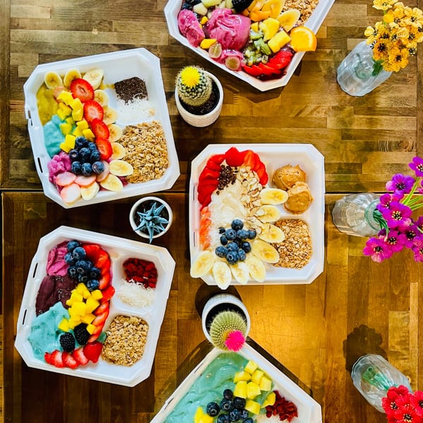Overhead view of five colorful acai bowls with various fruit toppings, granola, and nuts on a wooden table.