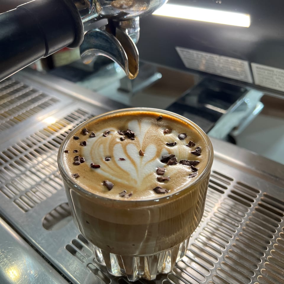 A latte with heart-shaped foam art and chocolate shavings sits on an espresso machine tray.
