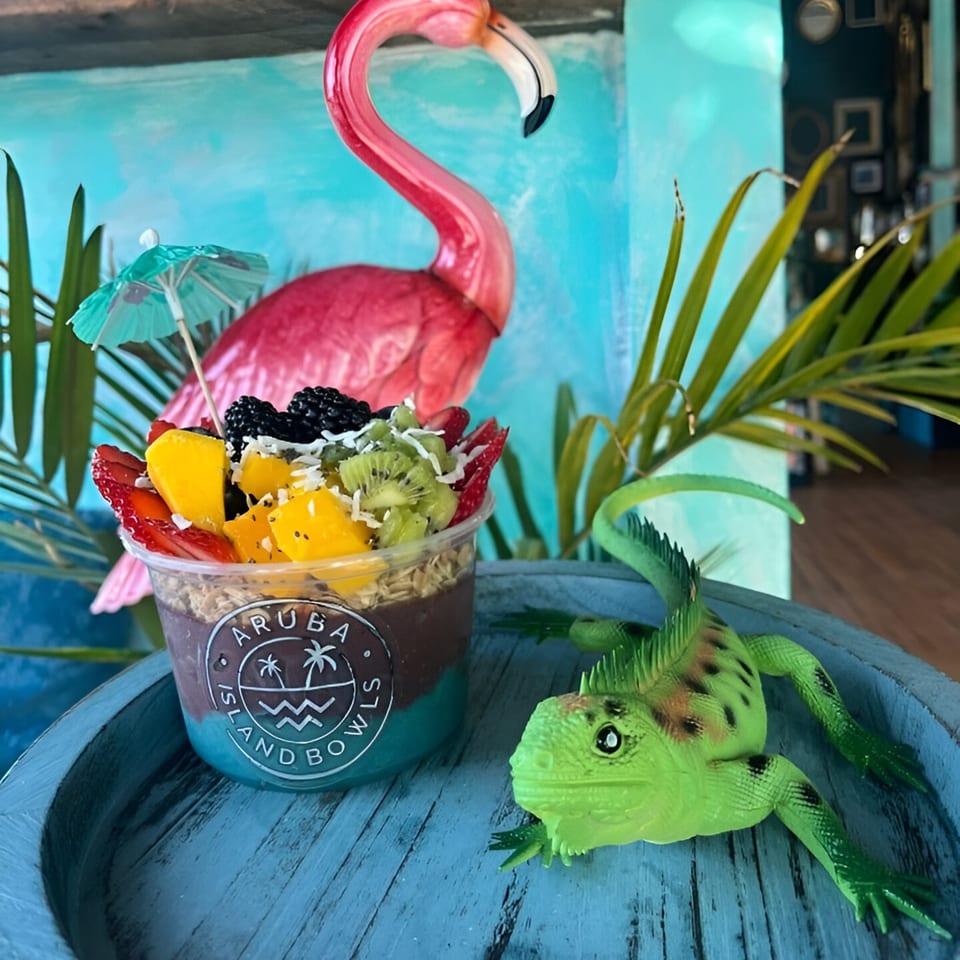 An Aruba Island Bowls acai bowl with fruit, a flamingo, and a lizard on a blue wooden surface.