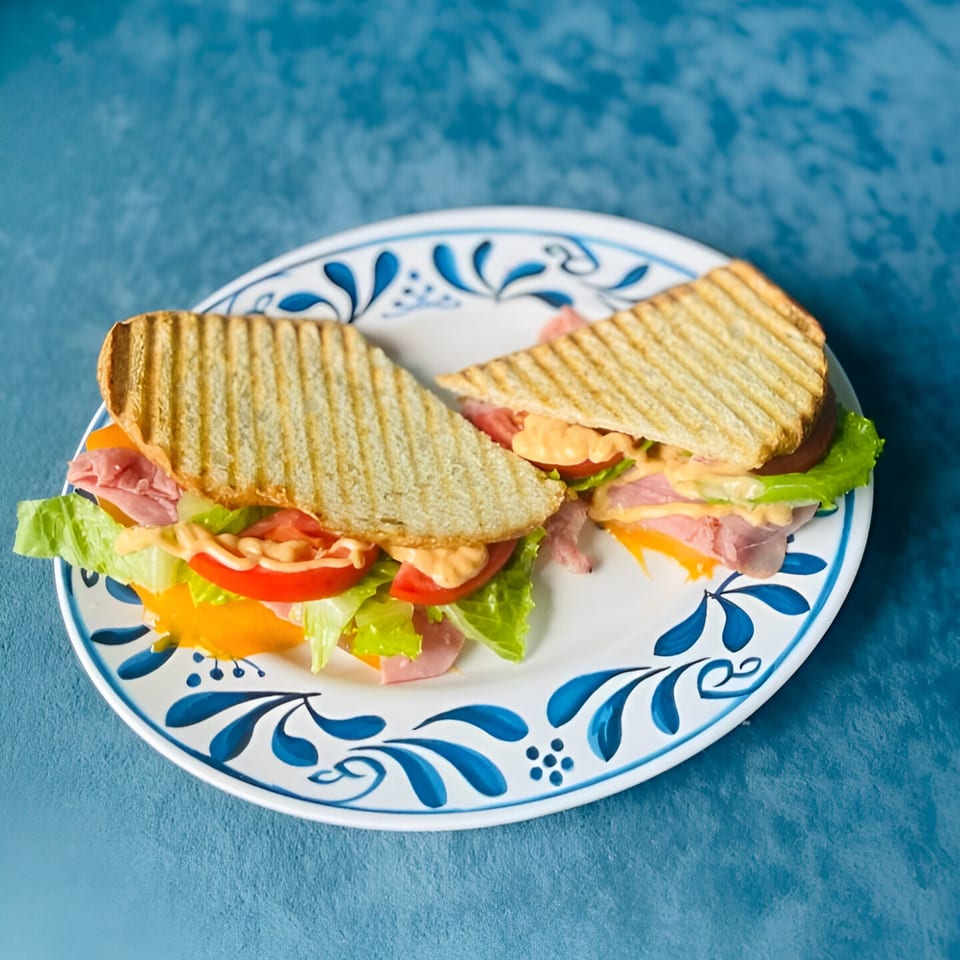 Two halves of a grilled sandwich with ham, cheese, lettuce, tomato, and sauce on a decorative plate.