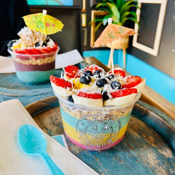 Two colorful Aruba Island Bowls with fruit, granola, and tiny umbrellas on a rustic blue table.