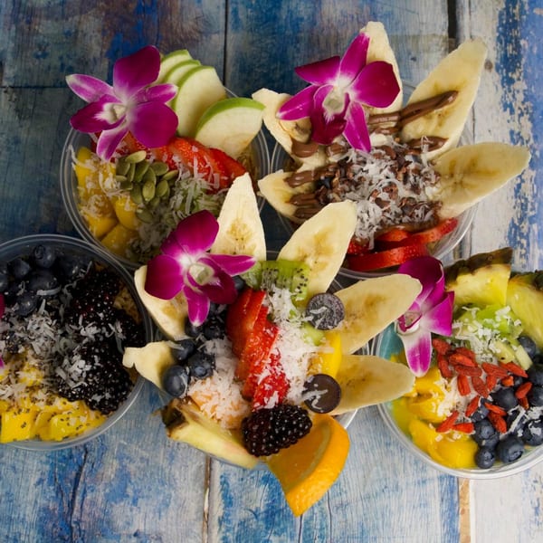 Five colorful acai bowls, garnished with fresh fruit, nuts, shredded coconut, and orchids on a blue wooden table.