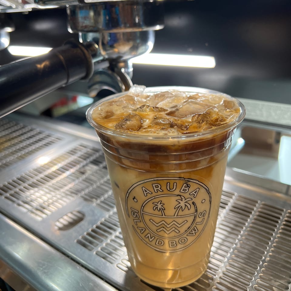 Iced coffee with the Aruba Island Bowls logo on a clear cup, next to an espresso machine.
