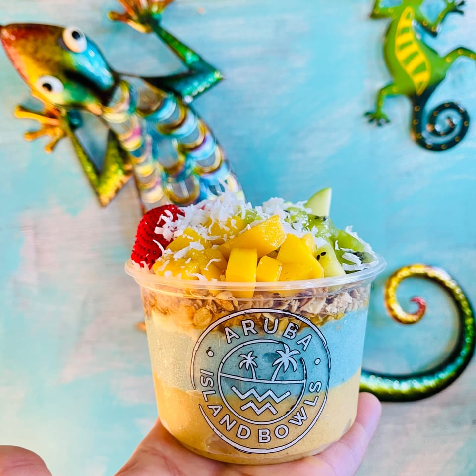 Hand holding an Aruba Island Bowls Cafe acai bowl with mango, kiwi, strawberry, and coconut.